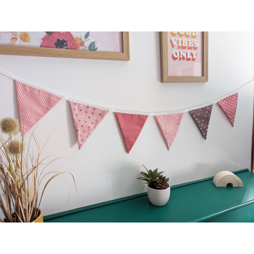 Guirlande décorative de fanions roses en tissu, suspendue sur un mur avec cadres et objets déco, idéale pour chambre d’enfant ou décoration intérieure.